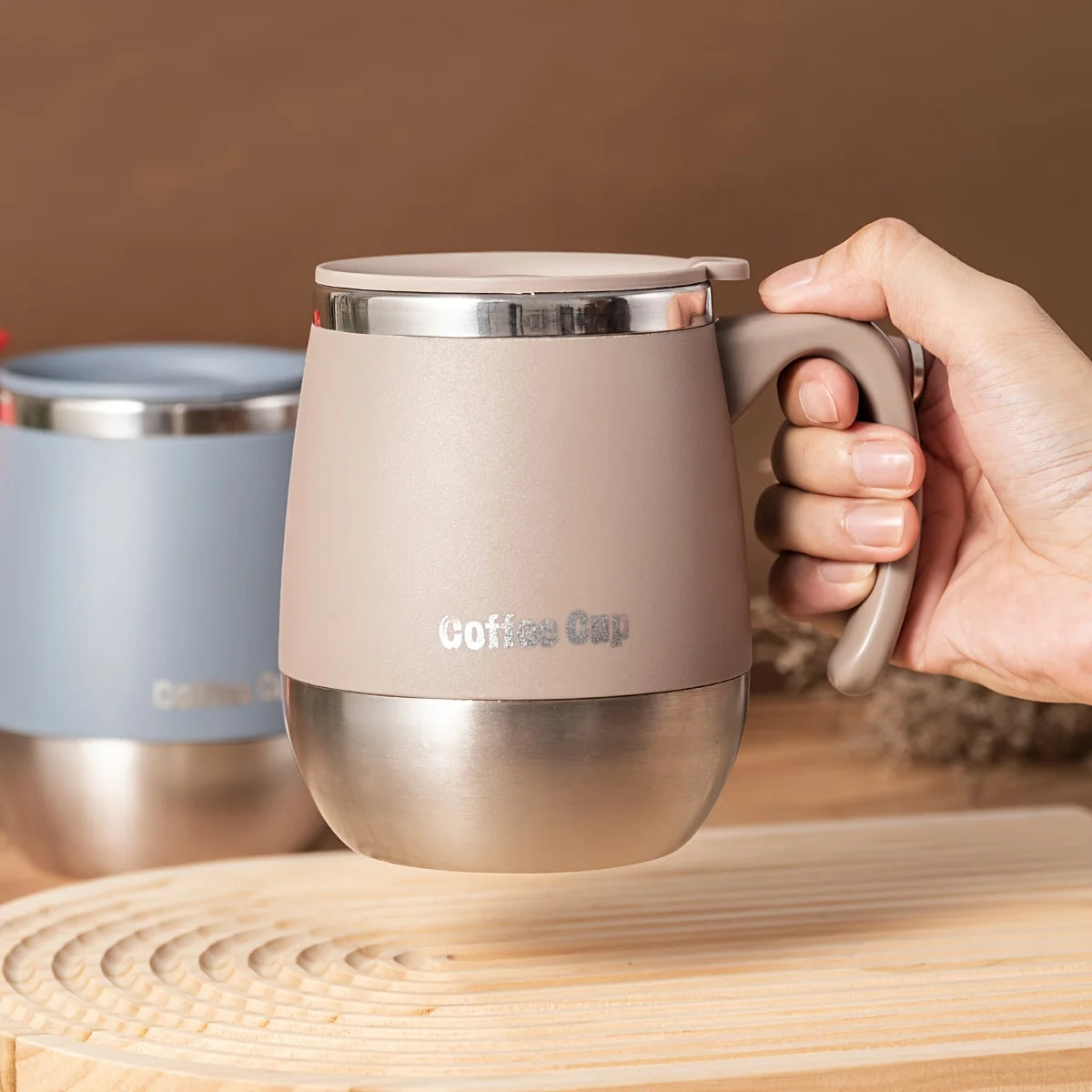 A hand holds a matte beige insulated stainless steel travel mug with a silver lid on a wooden surface.