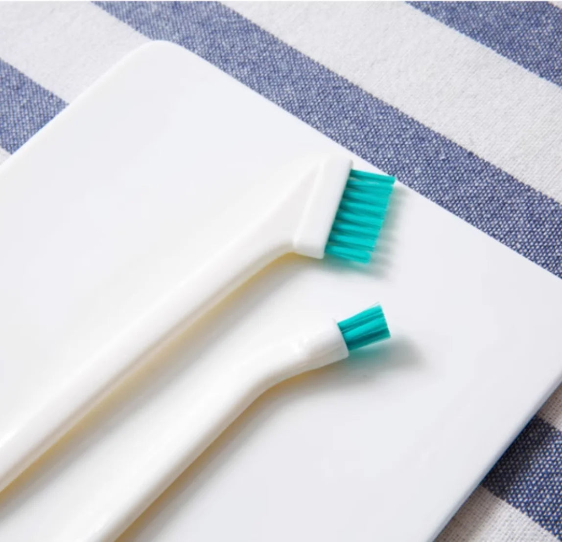 A white dual-sided silicone cleaning brush set with teal bristles resting on a striped cloth.