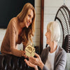 A woman is holding a mini wooden music box gift for another woman in a warm indoor setting.