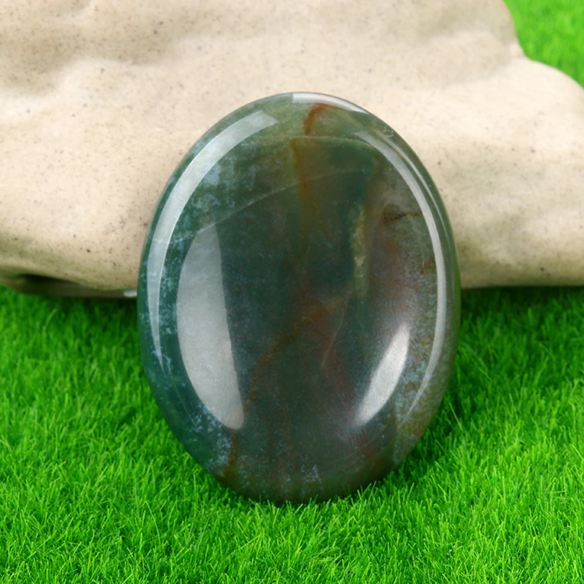 A polished green and brown natural quartz thumb stone resting on bright green grass.