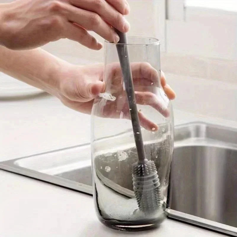 Hands are using a silicone bottle brush with a long handle to clean the inside of a glass bottle.