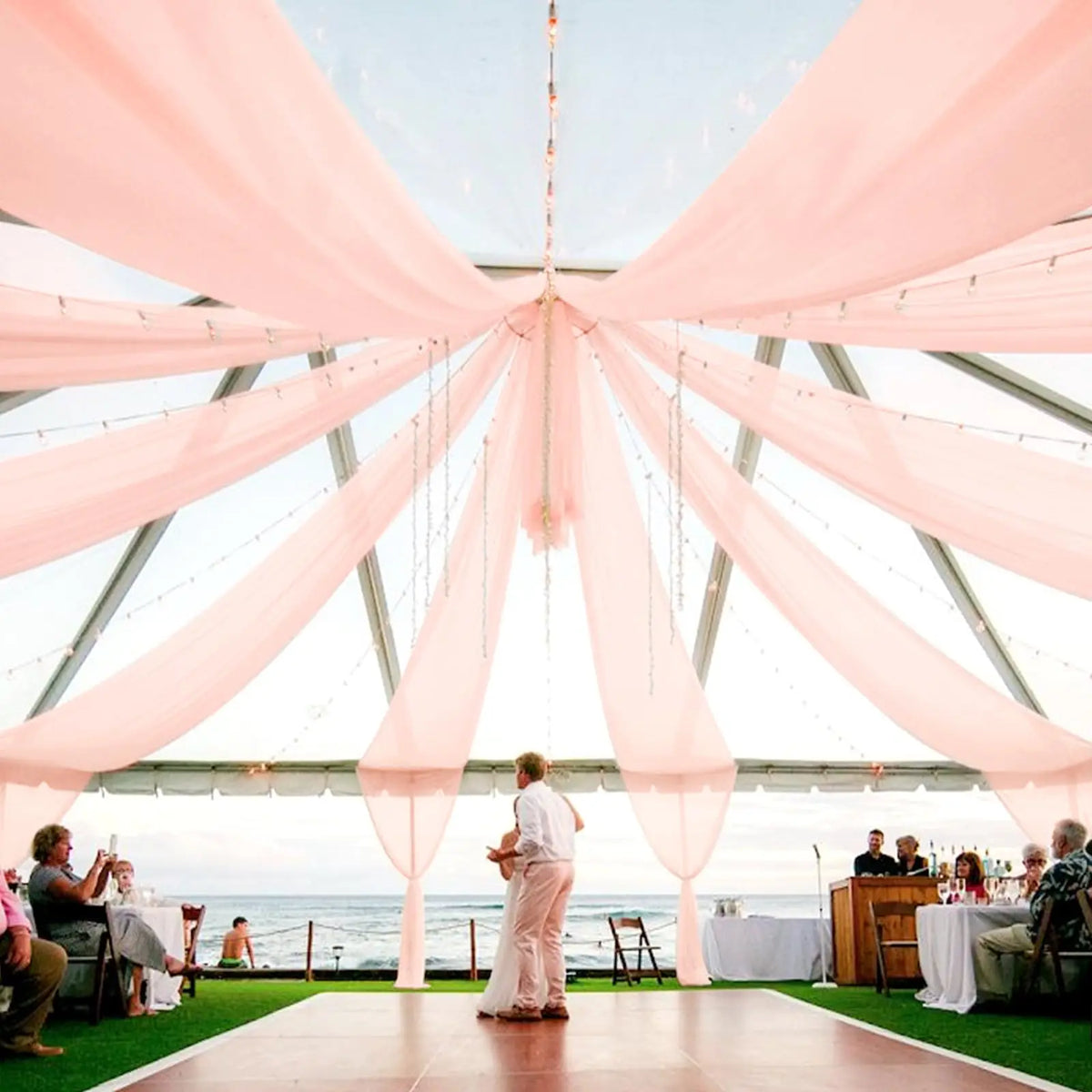A wedding or event setup featuring sheer pink chiffon draping fabric suspended from a metal structure over an outdoor area.