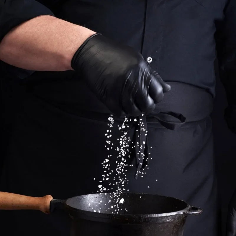 A person wearing black gloves is sprinkling white granular material into a dark pot.