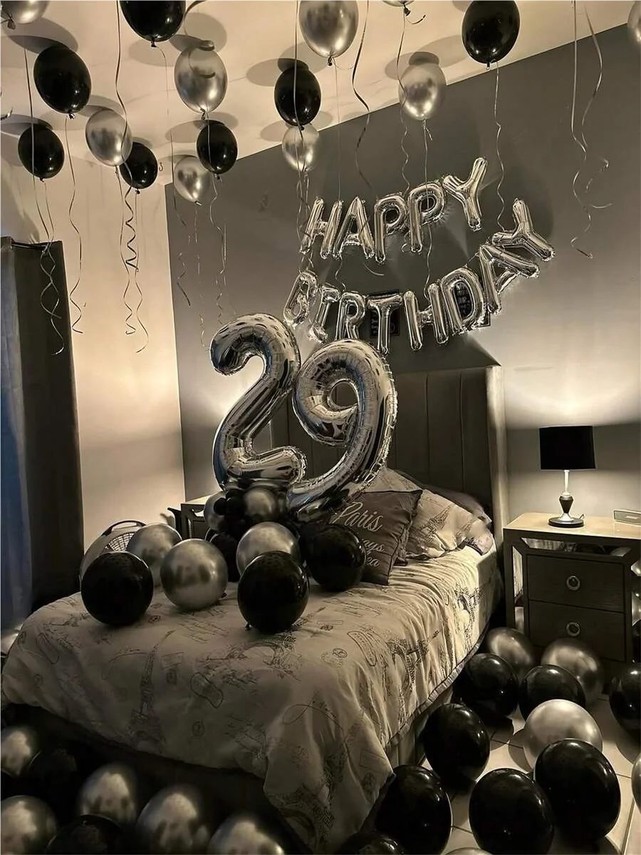 Decoración de cumpleaños con guirnalda de globos foil plateados y negros con el número 29 colgada sobre una cama.