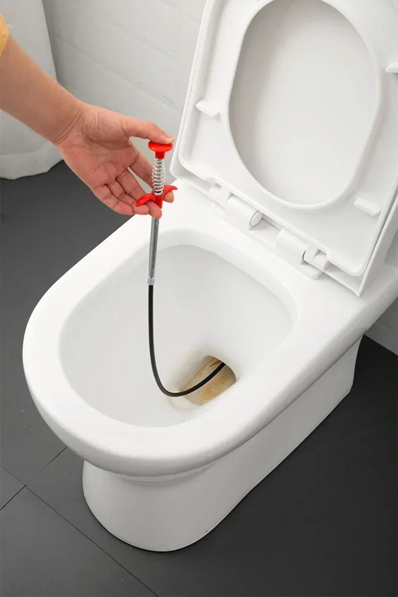 A hand is using a red and black sink and drain unclogging tool to clear a clog in a white toilet bowl.