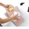 Hands place a yellow bar of soap and a white cloth onto a pink thickened washboard in a bathtub.