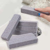 A hand is holding a gray pumice stone used for stain removal, with several stones visible on a white surface.