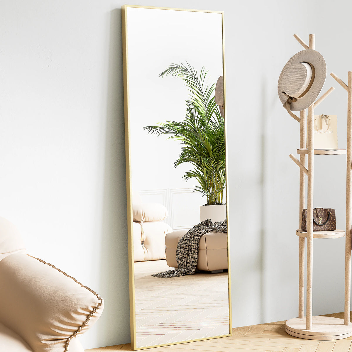 A full body standing wall mirror with a gold frame reflecting a green palm plant and a beige sofa.