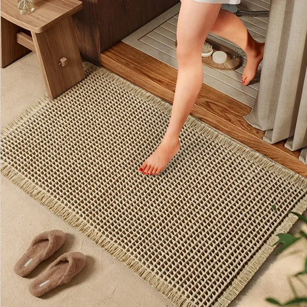 A beige waffle pattern bath mat is placed on a wooden floor next to a wooden stool and a pair of slippers.