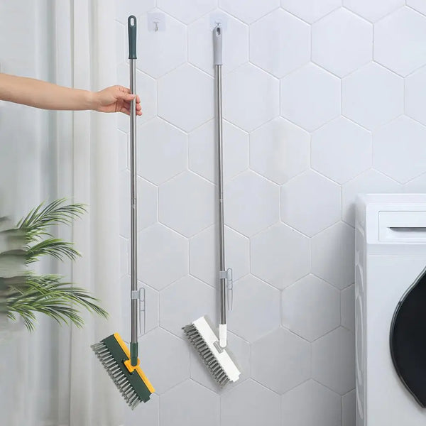 A long, silver floor scrub brush with a green head and a squeegee is mounted on a white tiled wall.