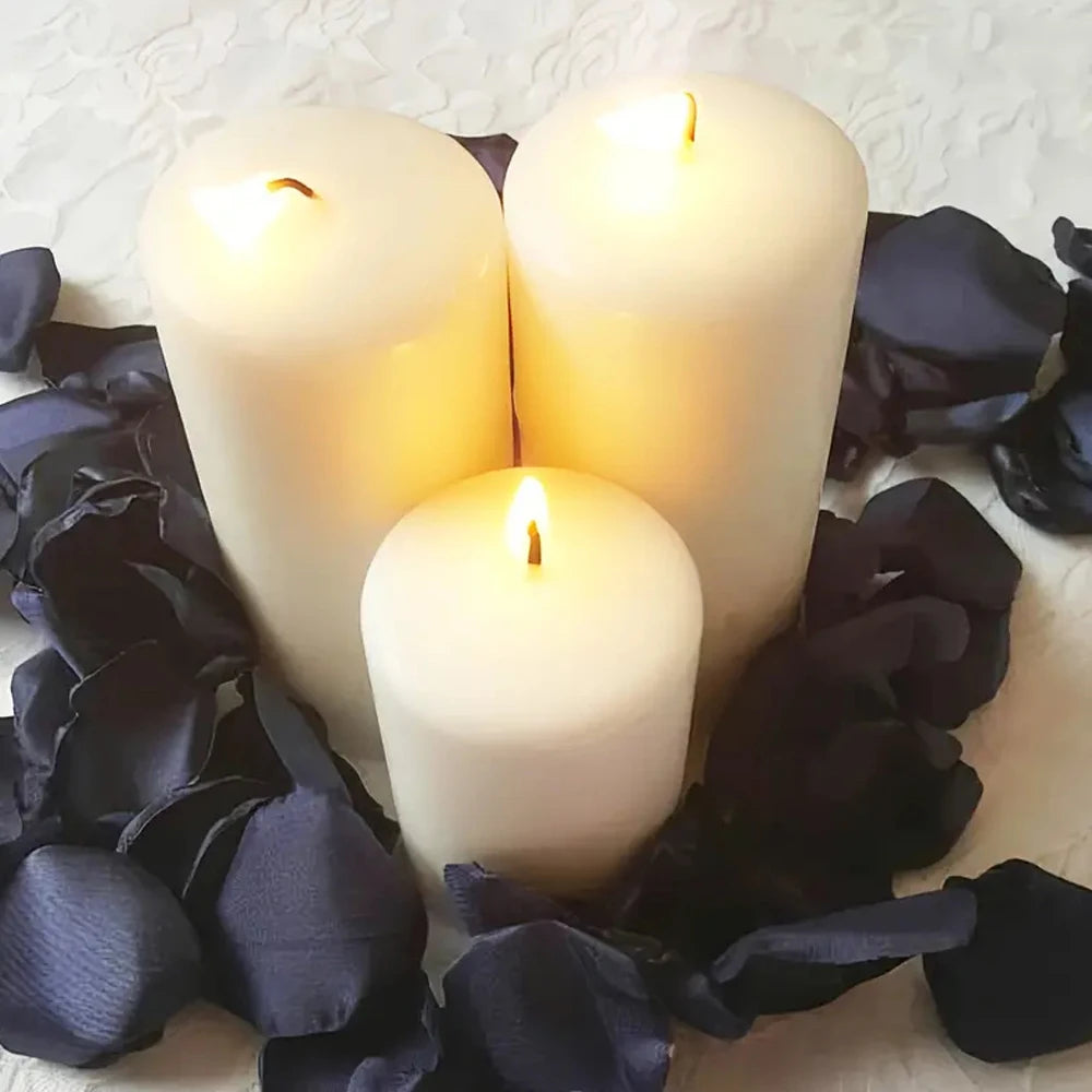 Tres velas blancas en forma de pilar rodeadas de pétalos de rosa de seda oscura sobre una superficie blanca.