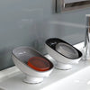 A white bathroom soap dish drainer with a leaf-shaped holder and an orange soap dish sitting on a white sink.