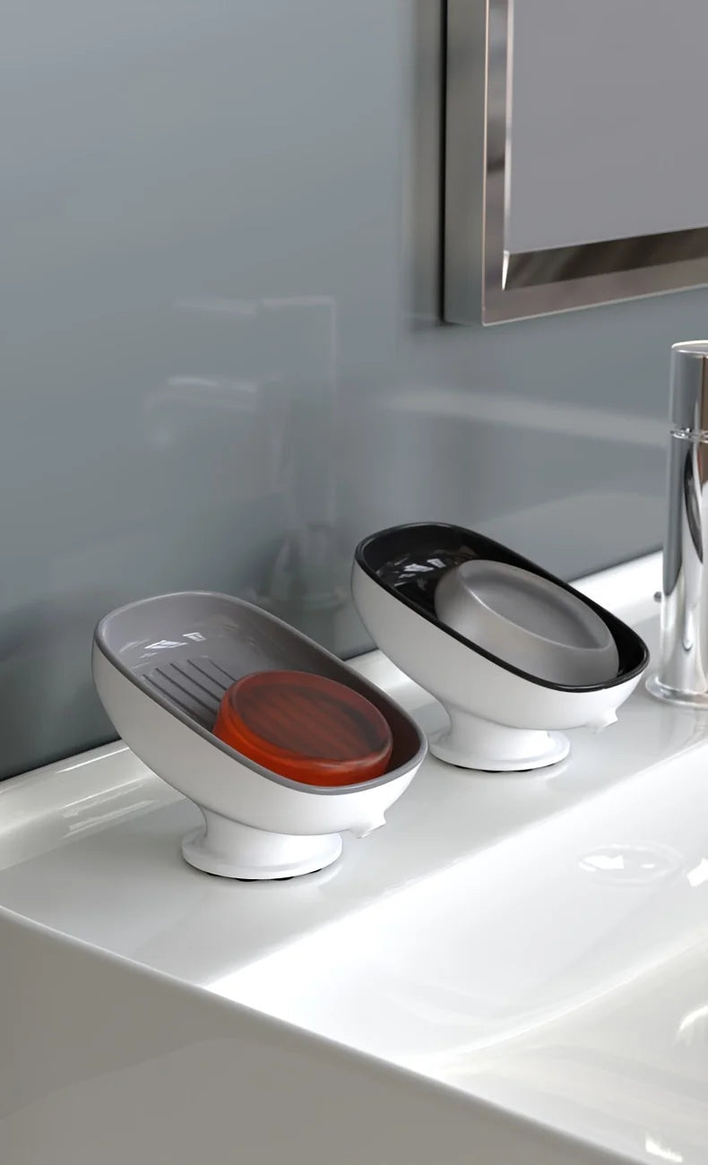 A white bathroom soap dish drainer with a leaf-shaped holder and an orange soap dish sitting on a white sink.