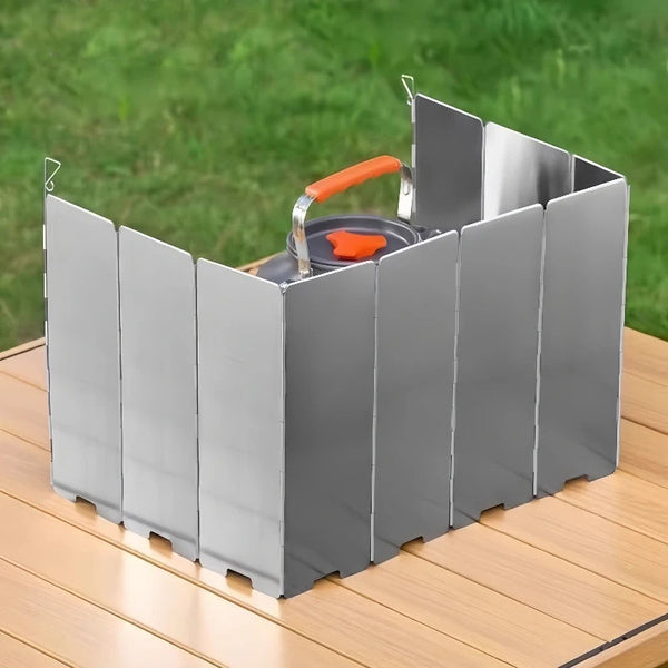 A silver aluminum windscreen stove shield with an orange handle is shown on a wooden surface.