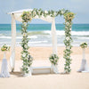 A wedding archway decorated with white fabric and garlands of artificial eucalyptus and roses on a sandy beach.