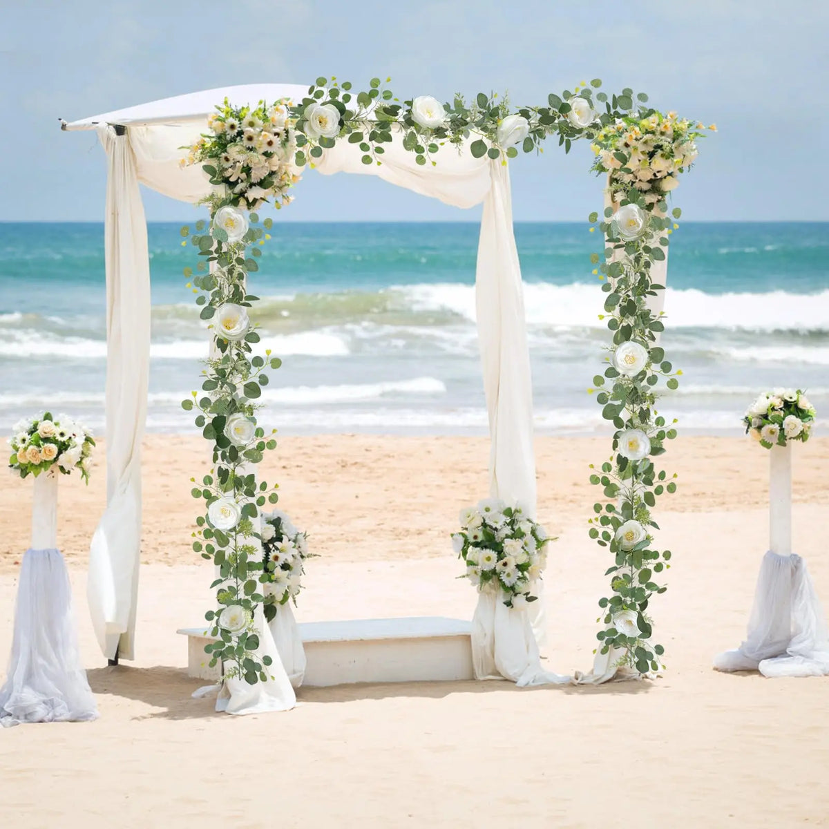 A wedding archway decorated with white fabric and garlands of artificial eucalyptus and roses on a sandy beach.
