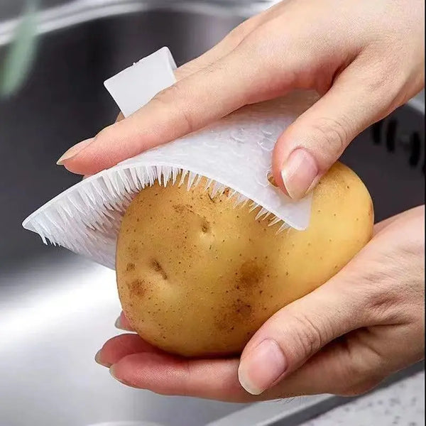 A hand is holding a yellow potato with a white silicone kitchen cleaning brush on it.
