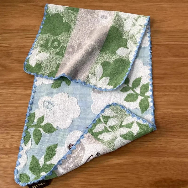 A folded square towel featuring a light blue background with white embroidered floral patterns and vertical stripes of green 