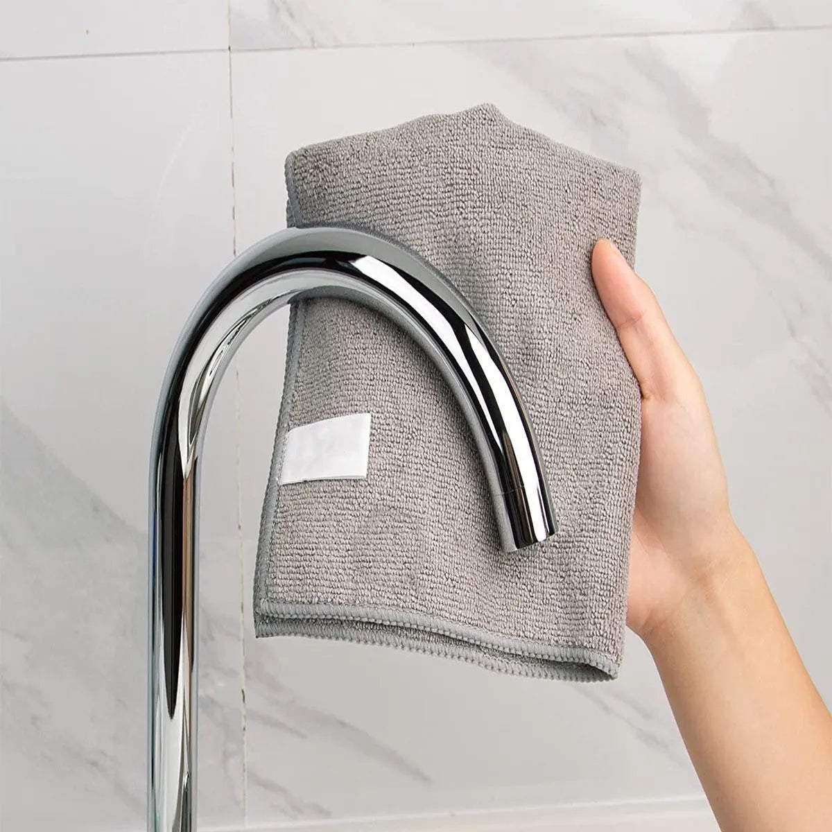 A hand is holding a gray microfiber cleaning cloth over a chrome faucet.