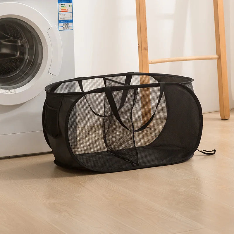 A black foldable mesh laundry basket is shown on a wooden floor next to a white washing machine.