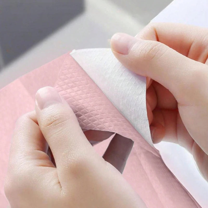 Eine Hand hält eine weiße, verbrauchsfertige Toilettendeckelabdeckung über ein rosa Papierprodukt.