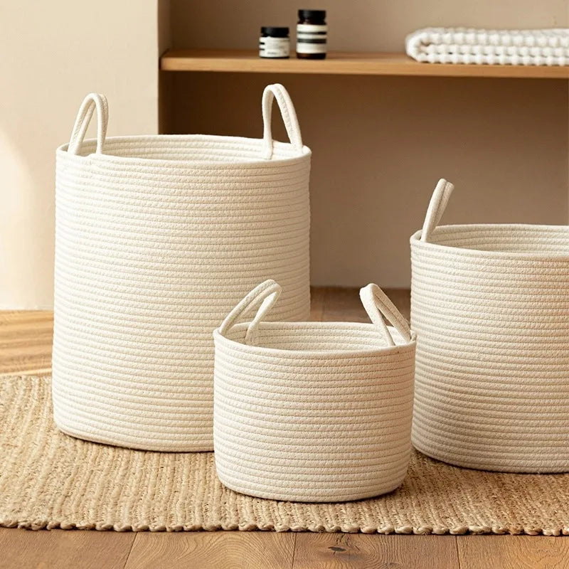 Three white natural cotton rope storage baskets sitting on a woven mat next to a wooden shelf.