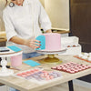 A person in a white coat is preparing pink frosting using reusable silicone piping bags and stainless steel tips on a wooden 