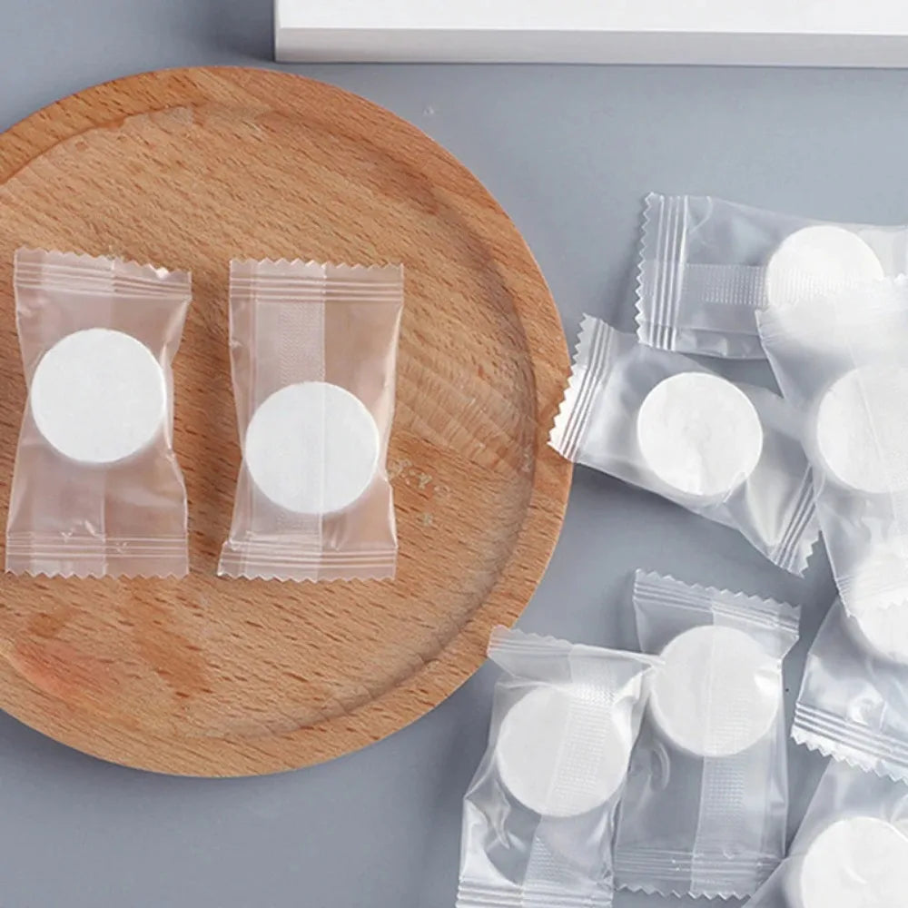 Several sealed packets of white disposable compressed travel towels are arranged on a wooden cutting board.