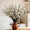 A vase filled with artificial white plum blossoms displayed against a white brick wall.