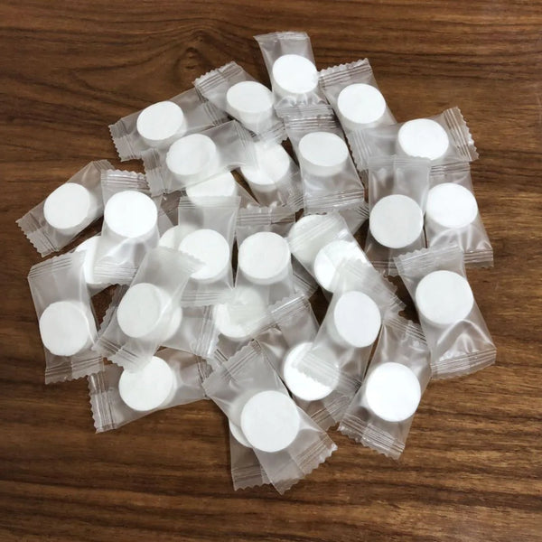 A pile of small, round white wipes packaged in clear plastic pouches on a wooden surface.