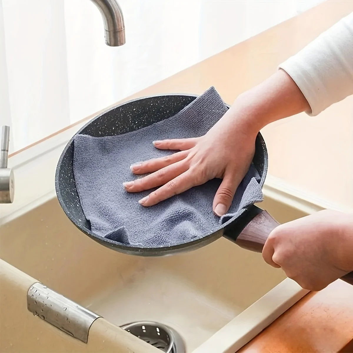 A hand is using a dark gray reusable multi-surface cleaning wipe on a kitchen sink.