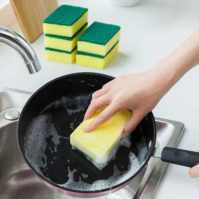 A hand is using a yellow scouring sponge to clean a black frying pan on a stovetop.