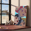 A vertical canvas art poster featuring stylized, abstract faces in muted pinks and blues displayed on a wooden shelf.