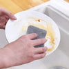 Hands are placing a gray double-sided cleaning sponge onto a white plate with some liquid residue on a kitchen sink.