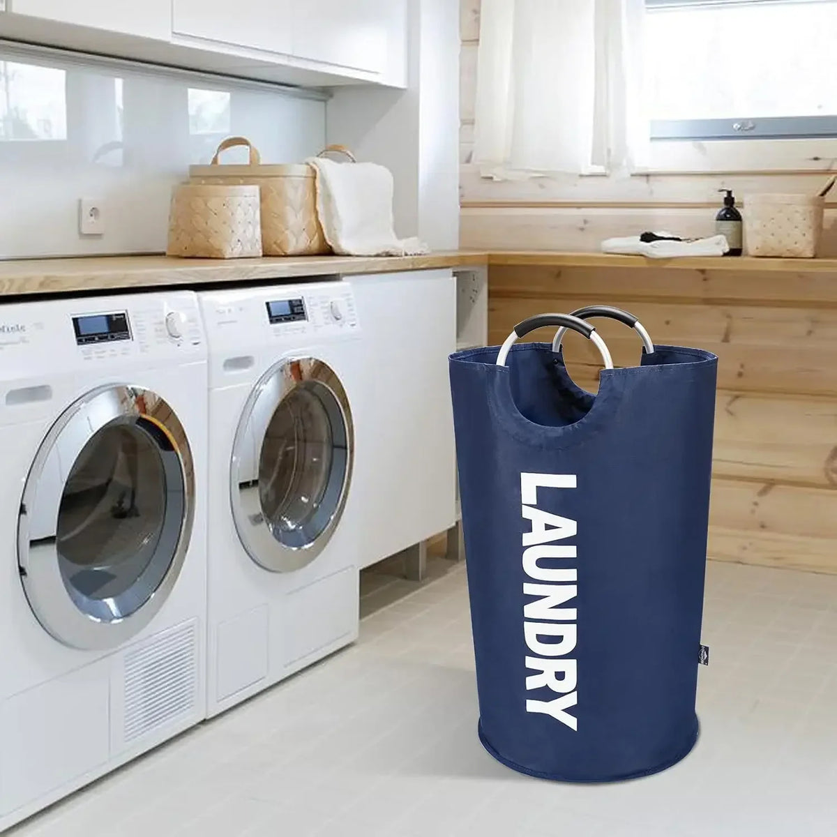 A dark blue foldable laundry basket with padded handles sits on a light floor in a laundry room.