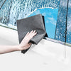 A hand holds a gray microfiber cleaning cloth against a window pane covered in water droplets.