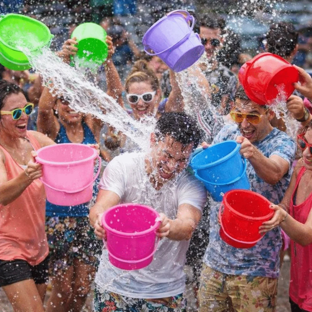 A group of people joyfully splashing water with six plastic buckets outdoors.