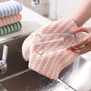 A hand is holding a clear glass and a peach-colored microfiber kitchen dish cloth on a stainless steel sink.