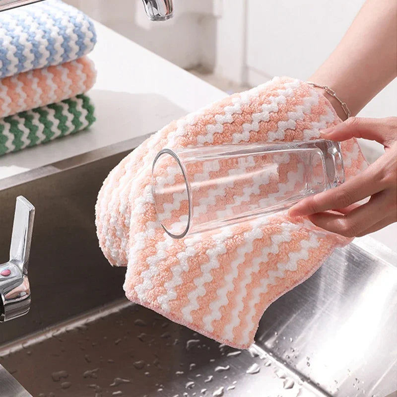 A hand is holding a clear glass and a peach-colored microfiber kitchen dish cloth on a stainless steel sink.