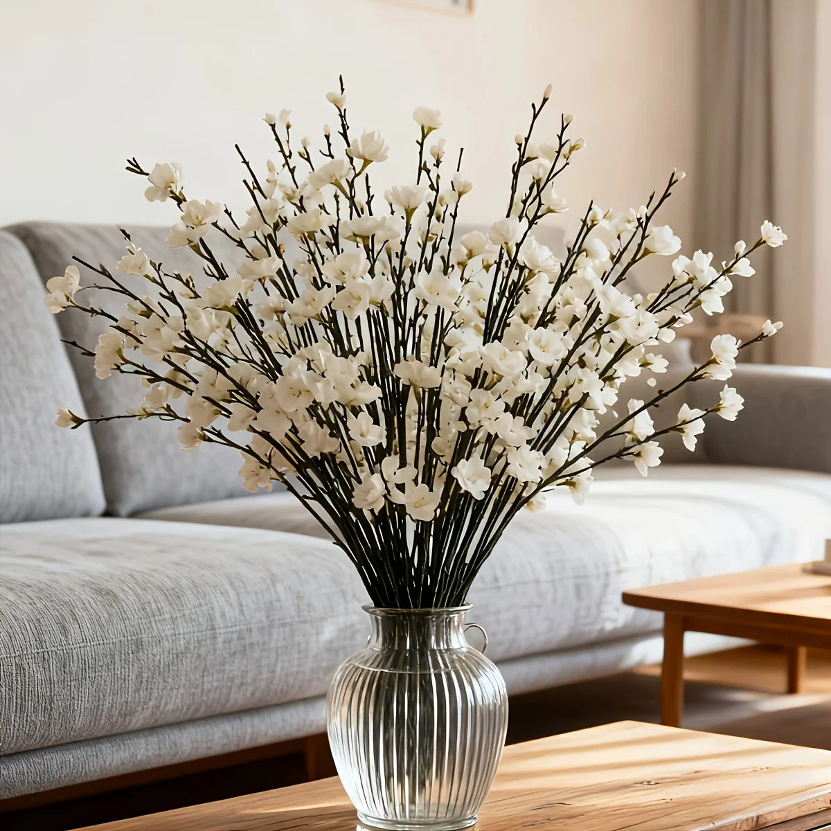 A vase filled with artificial white plum blossoms displayed on a wooden table next to a gray sofa.