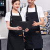 A man and a woman wearing black canvas aprons in a kitchen setting, holding an open notebook.