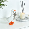 A white goose sculpture with orange feet and a metal key chain hanging next to a candle and black sticks.