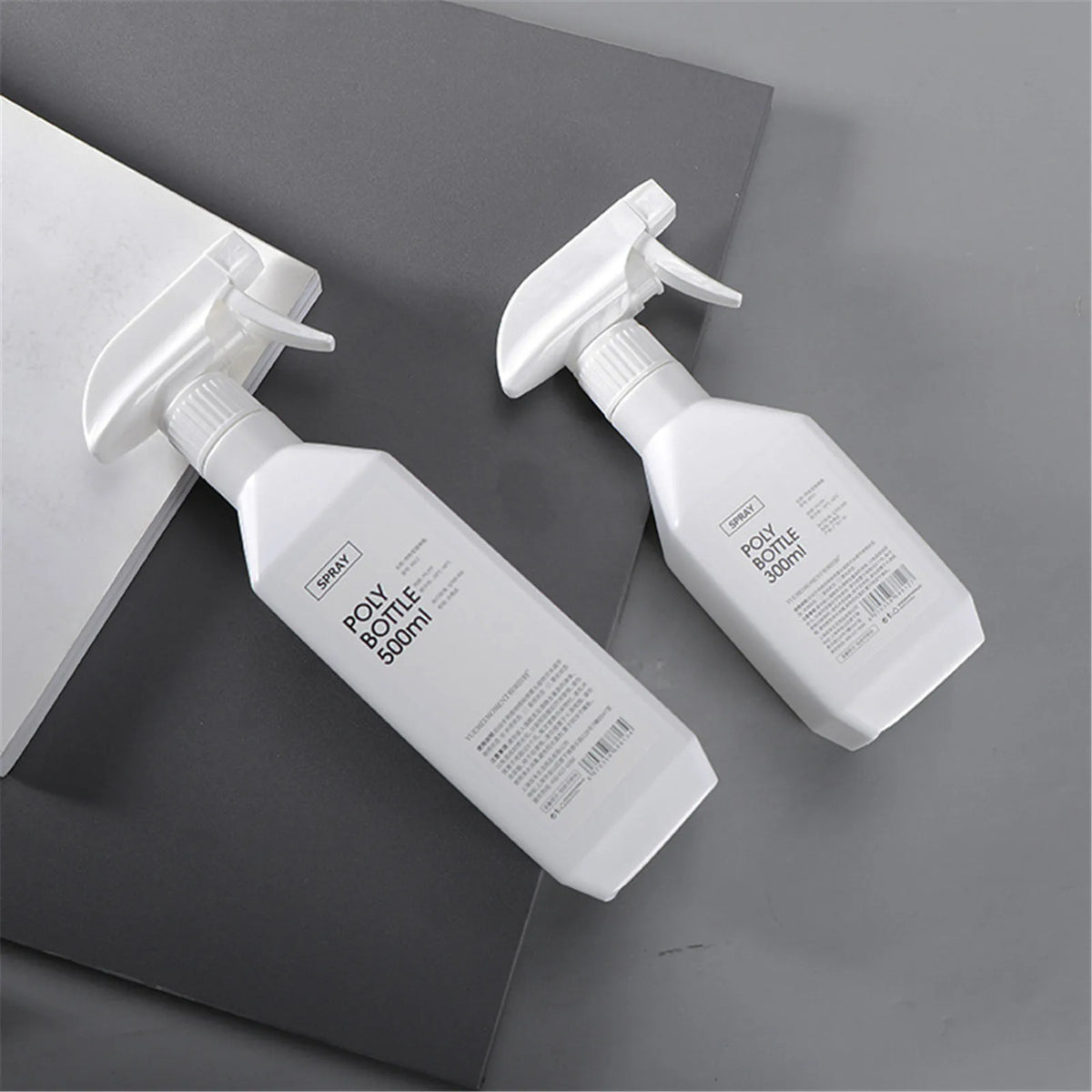 A white refillable plastic spray bottle with a pump dispenser is shown on a gray surface.