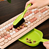 A hand uses a green handheld dustpan and brush set to clean a laptop keyboard.