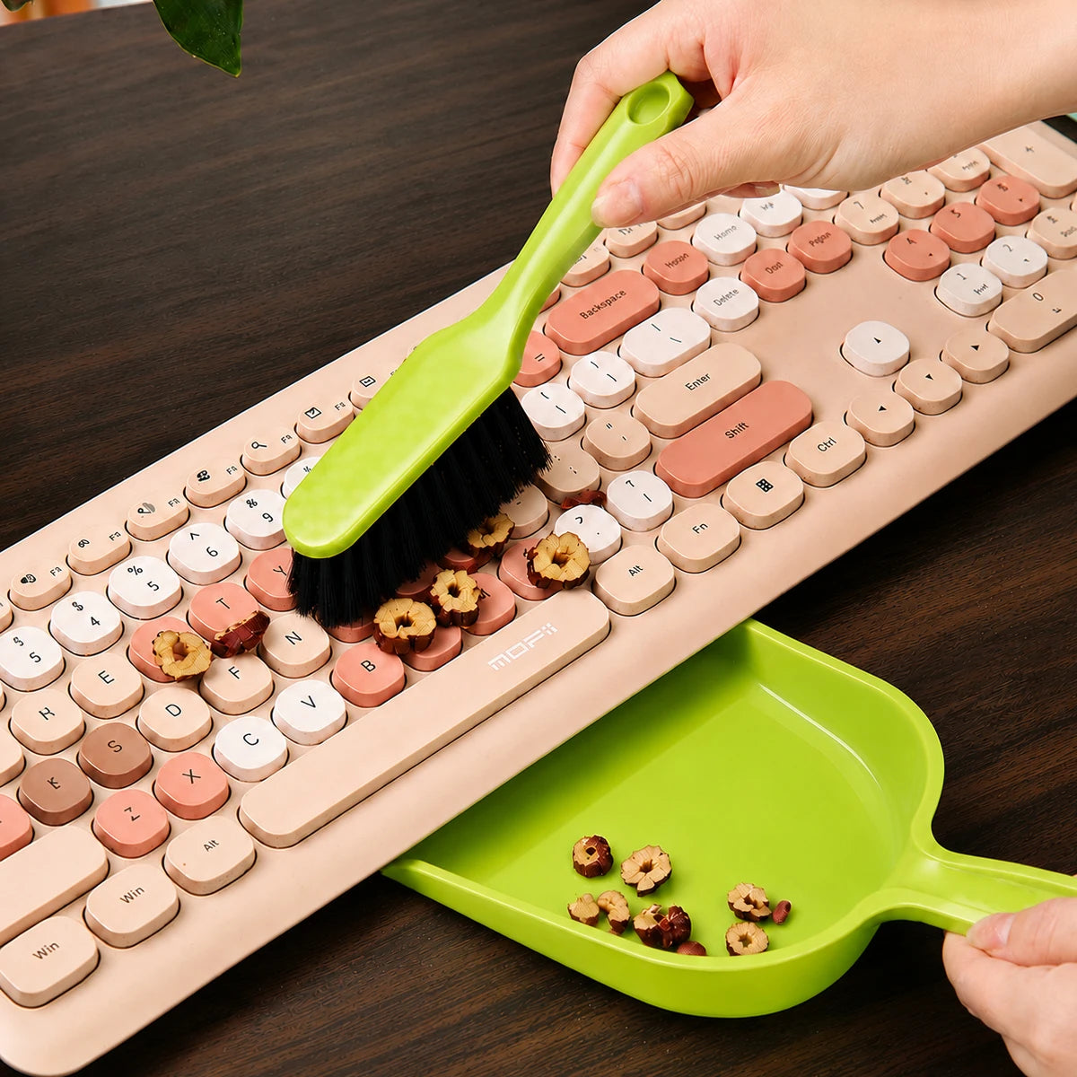 A hand is using a green handheld dustpan and brush set to clean a laptop keyboard.