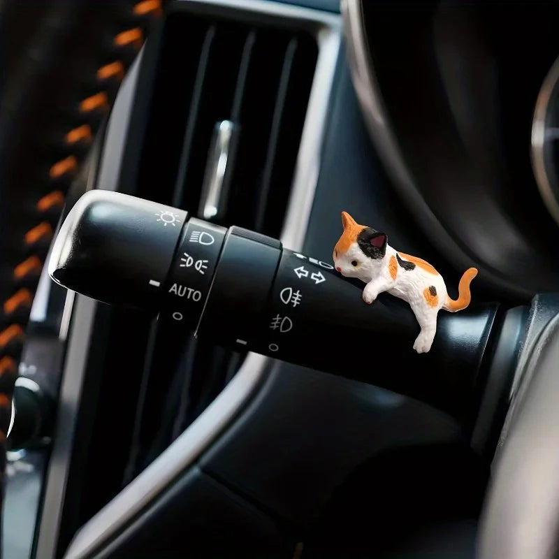 A small orange and white cat figurine is placed on a black control knob in a car interior.