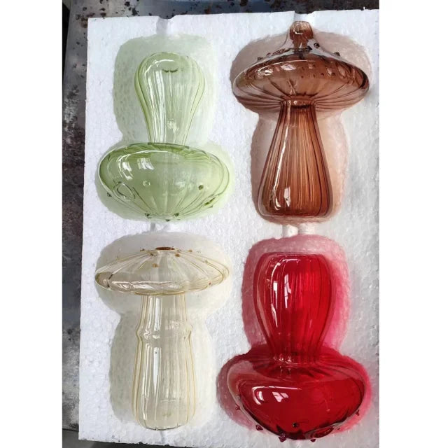 A collection of four Nordic style glass mushroom vases in shades of green, pink, and red displayed on white foam.