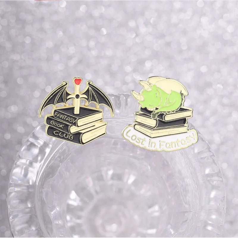 Pins de esmalte con temas de libros de fantasía, incluyendo un murciélago, una pila de libros y la cabeza de un dragón verde.
