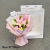 A bouquet of pink and yellow handmade crochet flowers wrapped in sheer paper placed inside a white paper shopping bag.