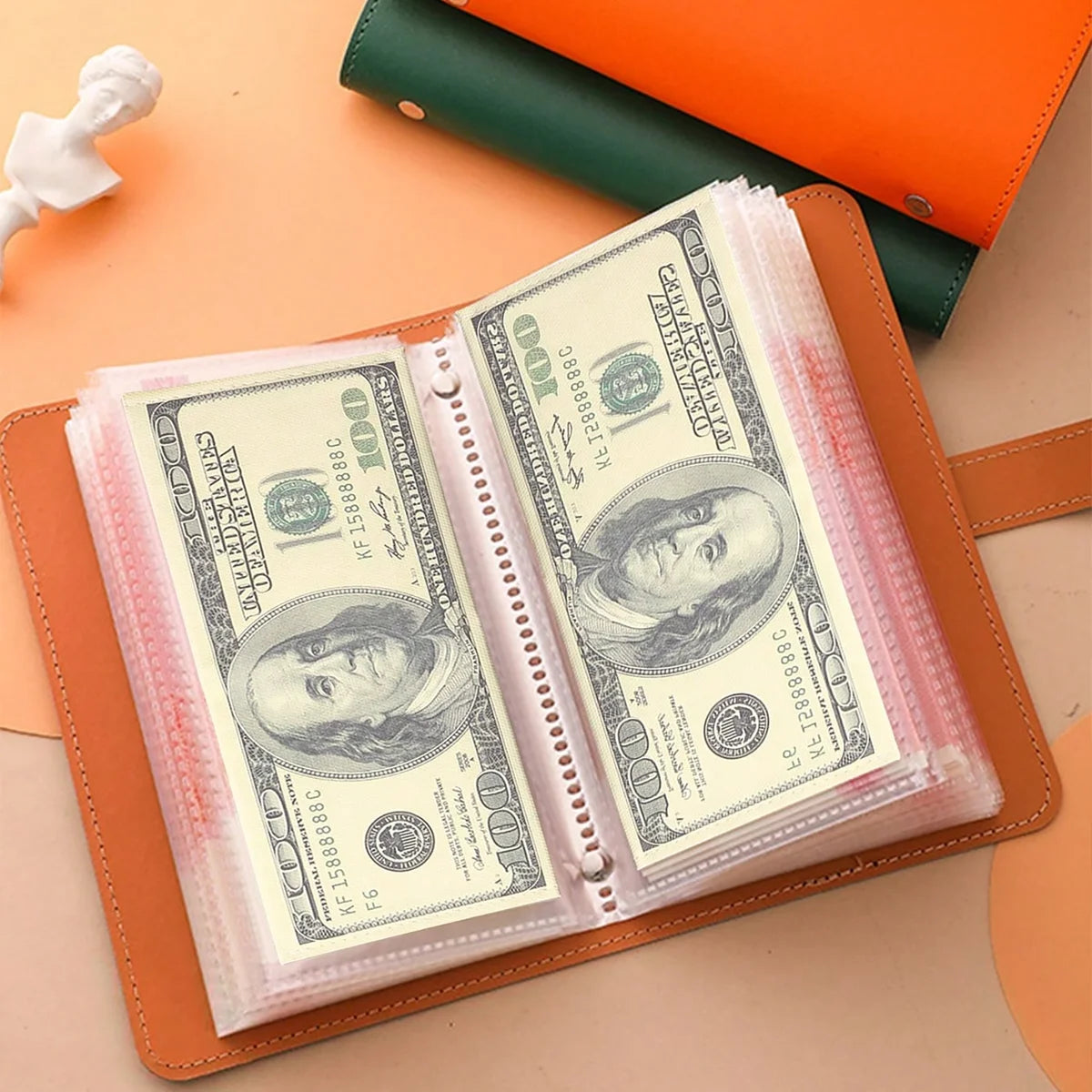 A leather cash organizer holds several hundred dollar bills in clear envelope sleeves on a light orange surface.